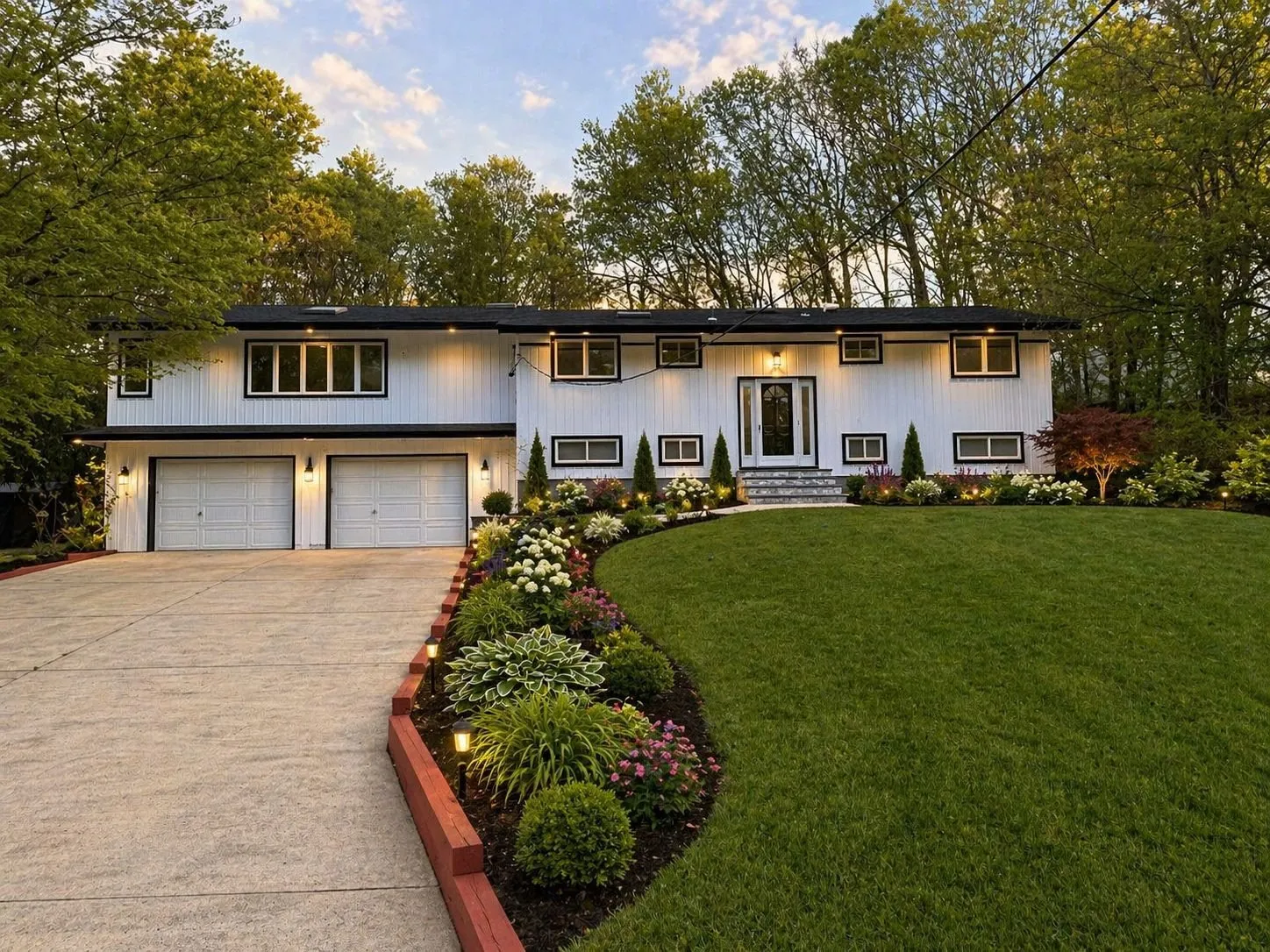 A Long Island front yard landscaped by Green Zone, with a lush lawn, fresh garden beds, and landscape lighting at dusk